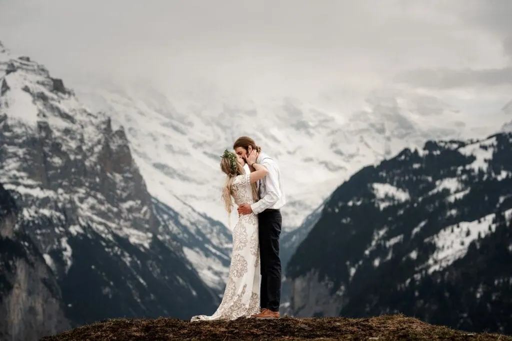 A bride and groom kiss on a hilltop during their Switzerland elopement, framed by snow-covered mountains in the background.