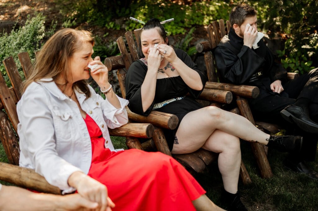 Three elopement guests outdoors sitting on wooden chairs, wiping away tears in an emotional moment during their friends elopement ceremony.