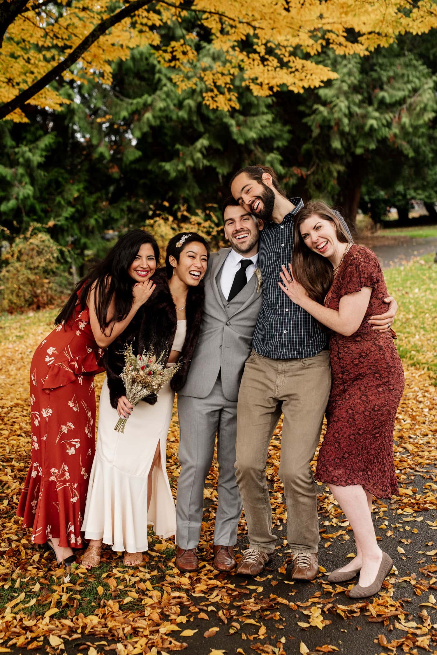 Five smiling people pose outdoors among autumn leaves, dressed in cozy clothing and surrounded by vibrant fall colors.