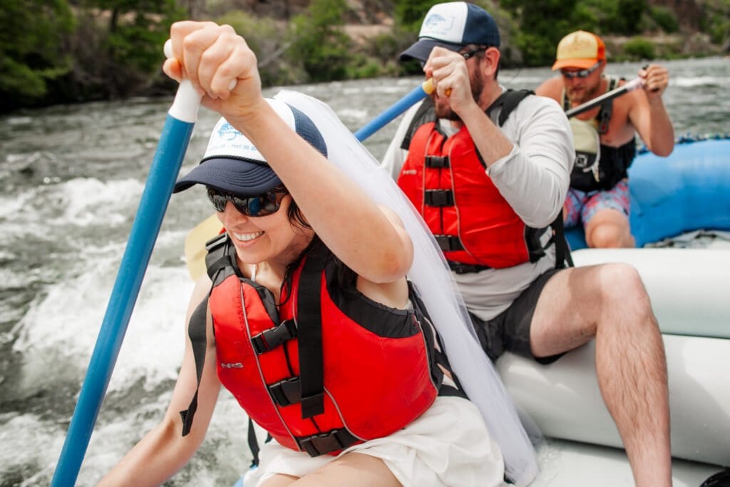 Three people wearing life vests paddle a raft down a river, led by a smiling bride at the front taking charge of the adventurous elopement.