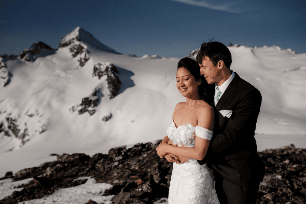 A bride and groom embrace on a rocky, snow-covered mountain peak during a Northern Cascades elopement.