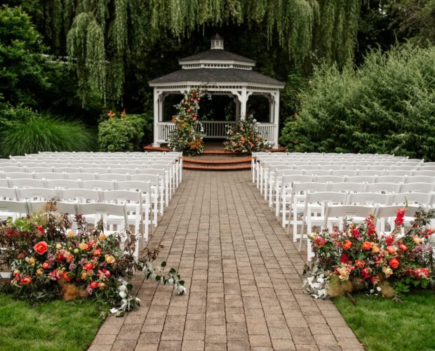 An outdoor wedding setup at Abigails Garden, Abernethy Center, features a white gazebo surrounded by greenery, a willow tree, and colorful flowers lining the aisle in Portland, Oregon.