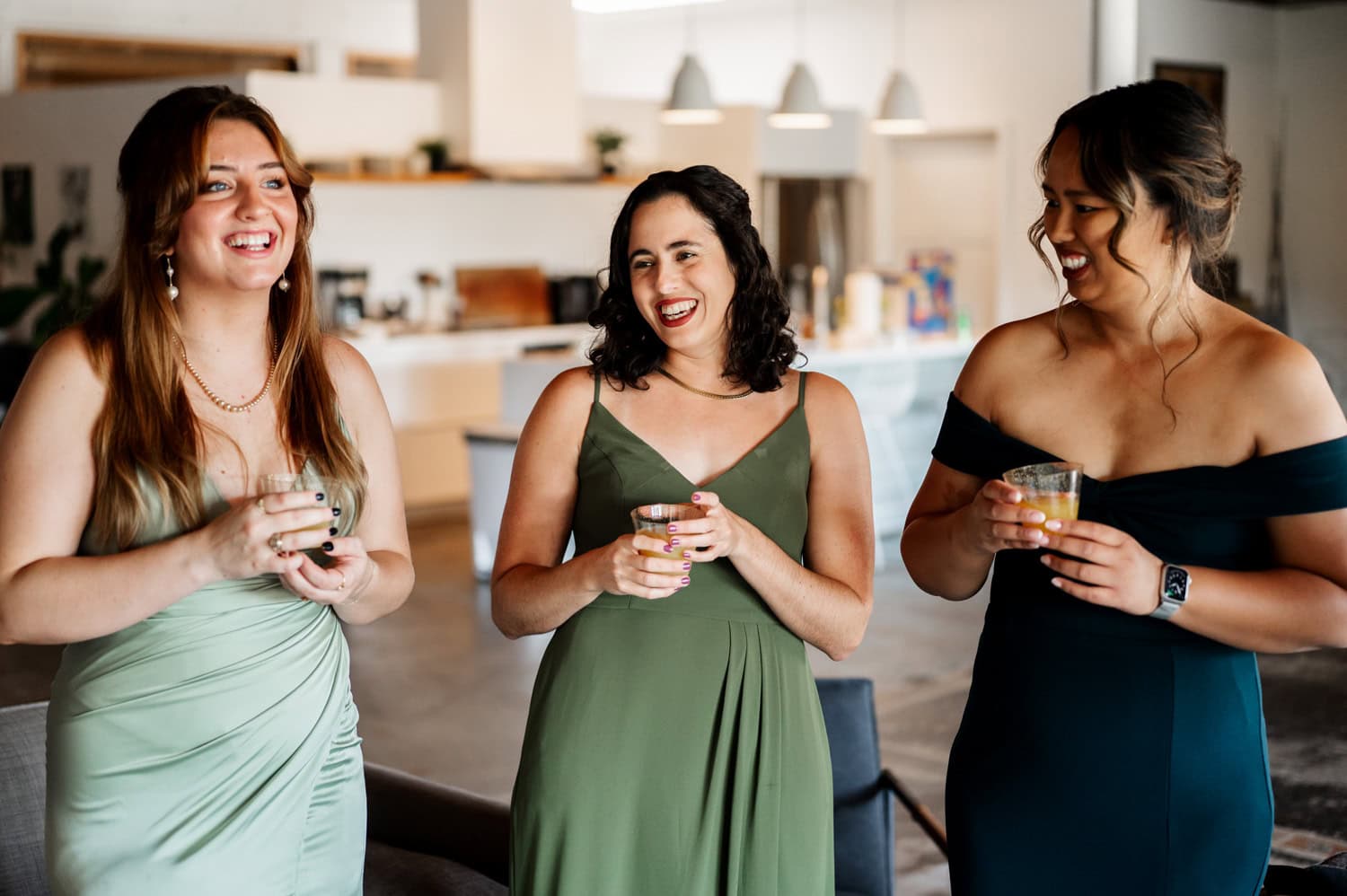Three women in elegant dresses laugh and hold drinks in a modern, well-lit kitchen, celebrating before a wedding at Union/Pine.