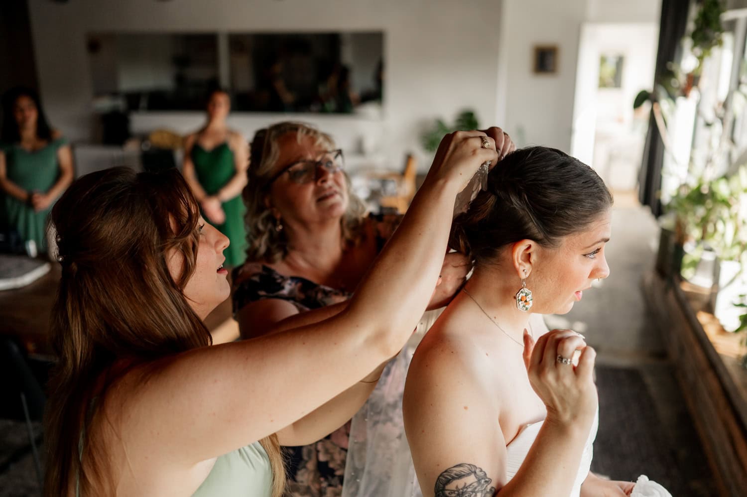 The bride is standing in a sunlit room, getting ready with assistance from two women. She wears a white dress, and the women help adjust her veil. The setting is Union/Pine, with light streaming through large windows, illuminating the scene.