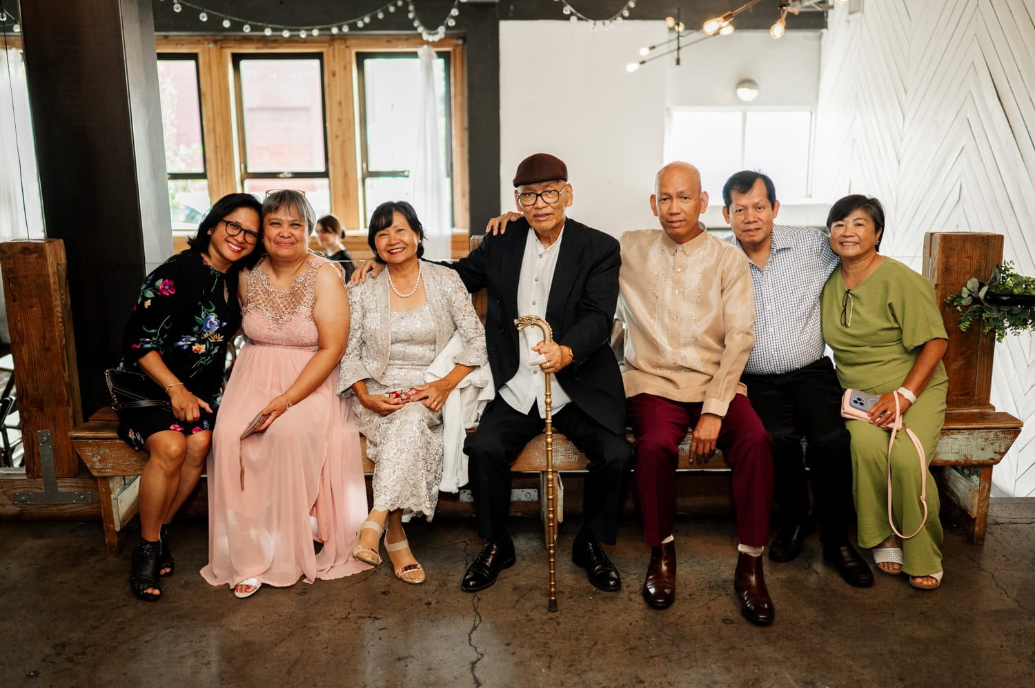 Seven people are seated on a bench indoors, dressed formally and smiling warmly at the camera. They convey a sense of joy, possibly celebrating a special occasion at Union/Pine.