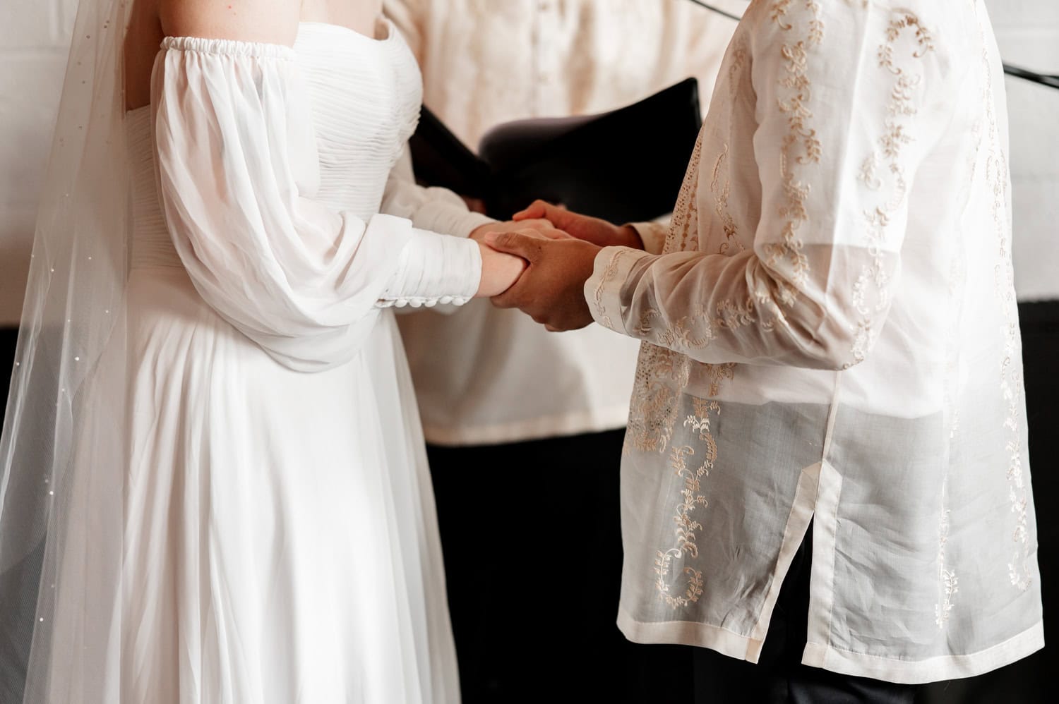 The bride in a white dress and groom in an embroidered shirt hold hands during their wedding ceremony at Union/Pine.