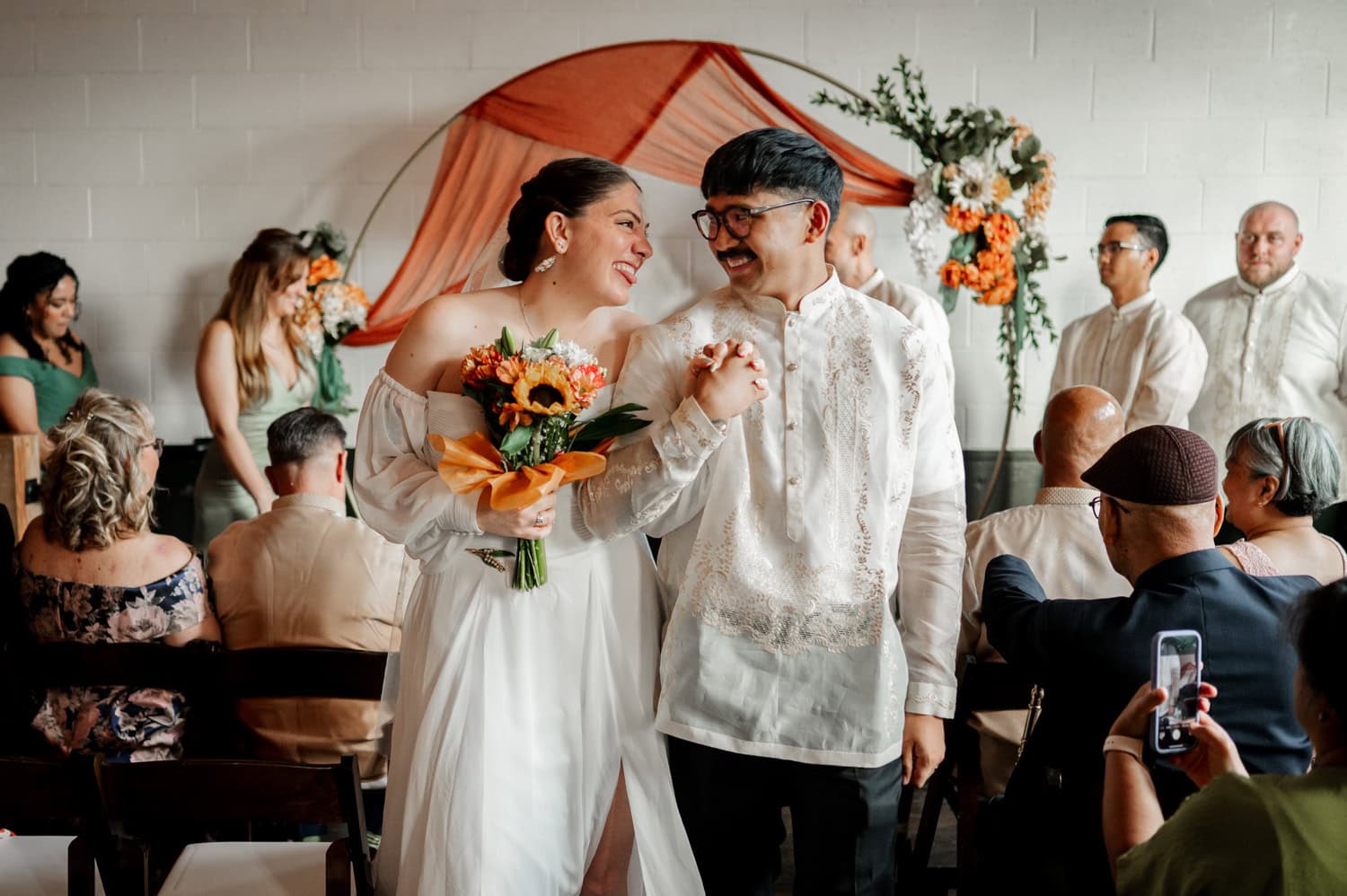 A joyful couple holds hands and smiles as they walk down the aisle at their wedding ceremony at Union/Pine, with delighted guests surrounding them.