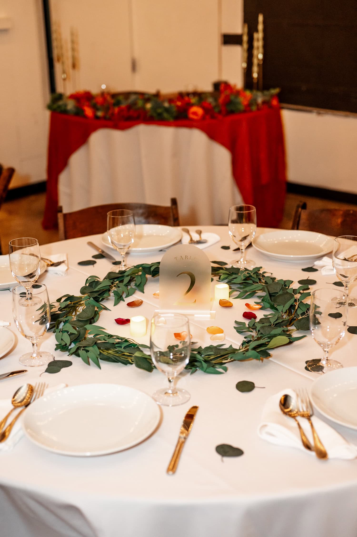 A round table covered with a white cloth is set for a wedding at Union/Pine, featuring plates, glasses, and cutlery. The centerpiece consists of greenery and candles, creating a warm and inviting atmosphere.