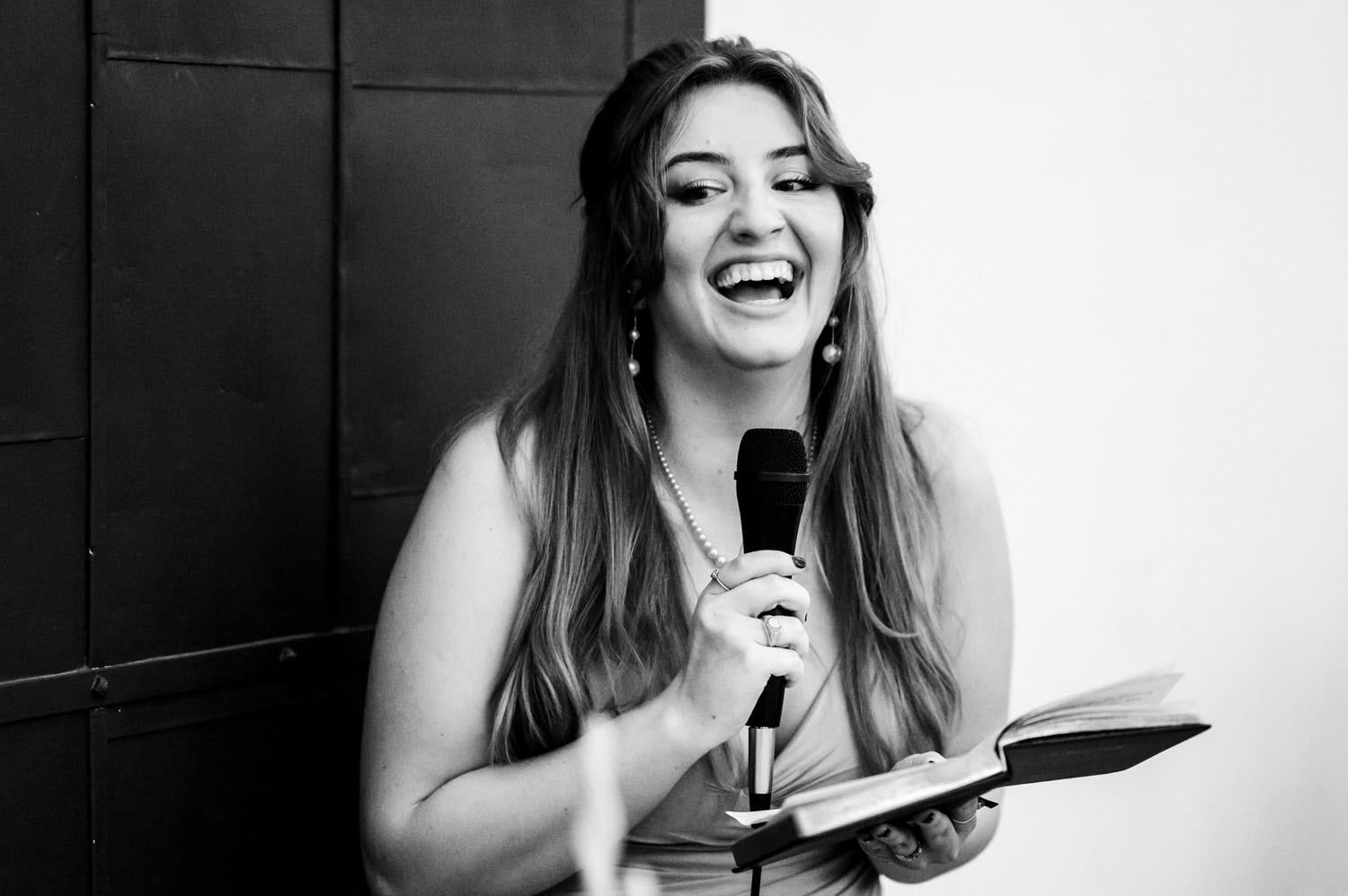A woman smiling warmly speaks into a microphone, holding an open book, in a black and white setting at a venue with the name Union/Pine.