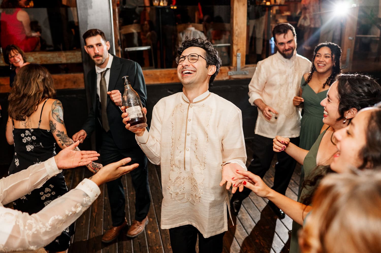 A joyful group of people dancing and laughing at a wedding celebration at Union/Pine, with one person holding a bottle.