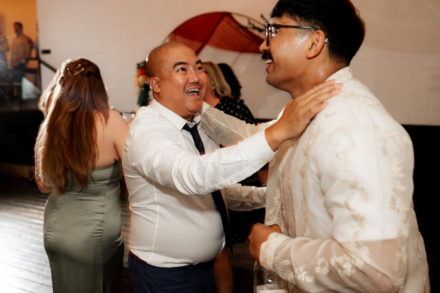 Two men smiling and embracing at a wedding, with a woman in a green dress standing in the background at Union/Pine.