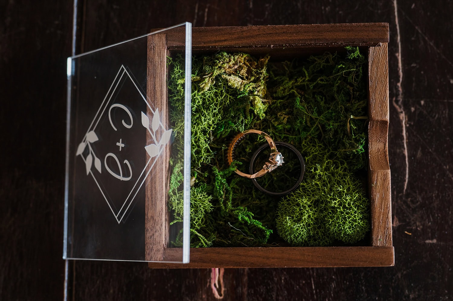 A wooden box with a moss interior holds wedding rings, with engraved initials on the glass lid.