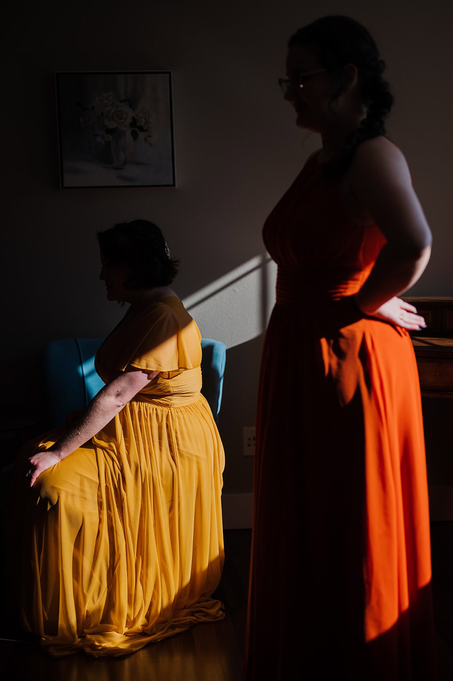 Two women in flowing yellow and orange dresses sit and stand against a dark background, their silhouettes elegantly highlighted by the light.