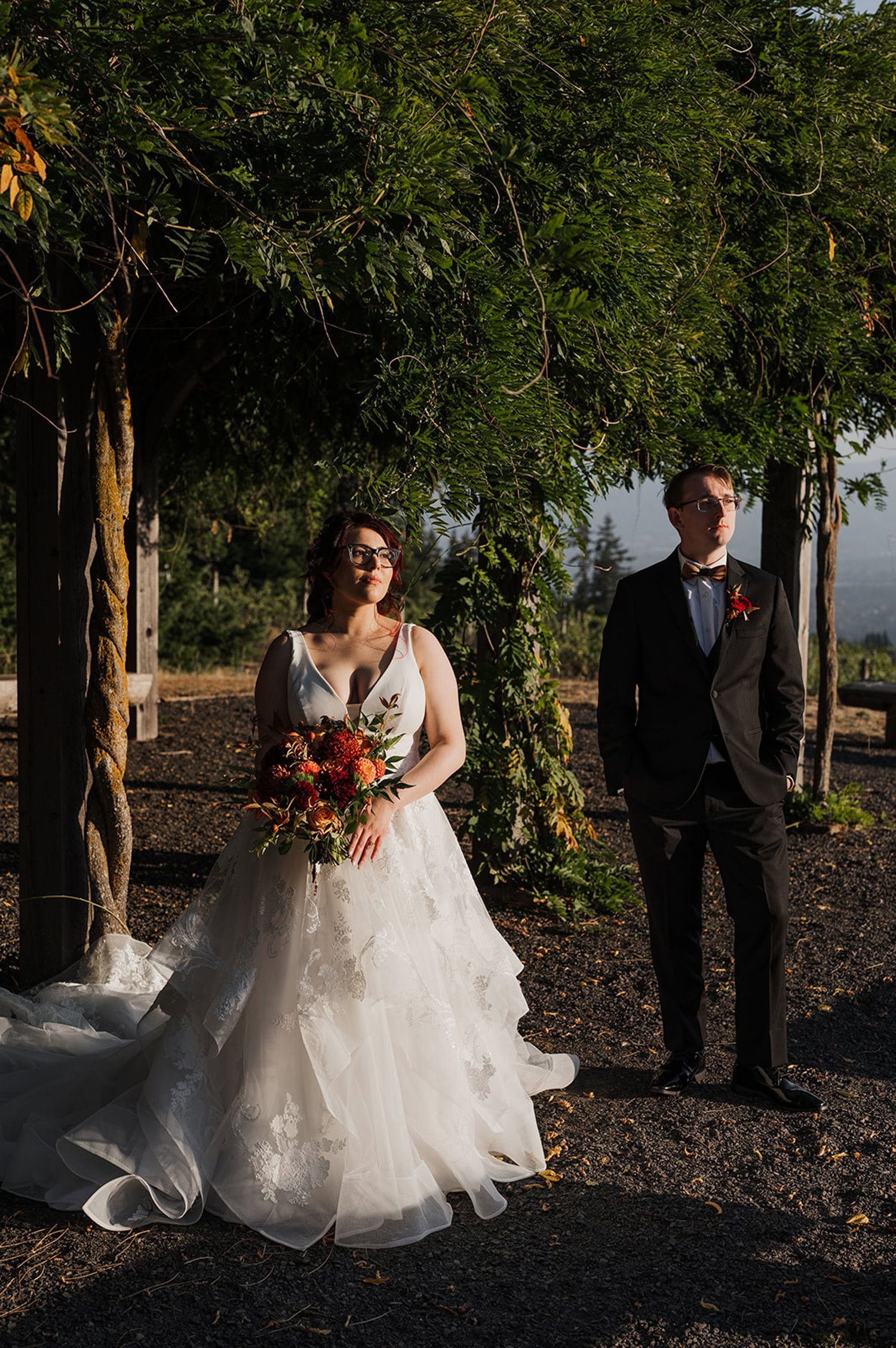 At Gorge Crest Vineyards, a bride in a flowing white gown stands with her groom in a dark suit under a lush green pergola, holding a bouquet.