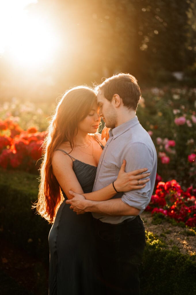 A woman with red hair wearing a green dress embraces her fiancé in a blue dress shirt with roses in the background and warm sunset light glowing from behind during their engagement session.