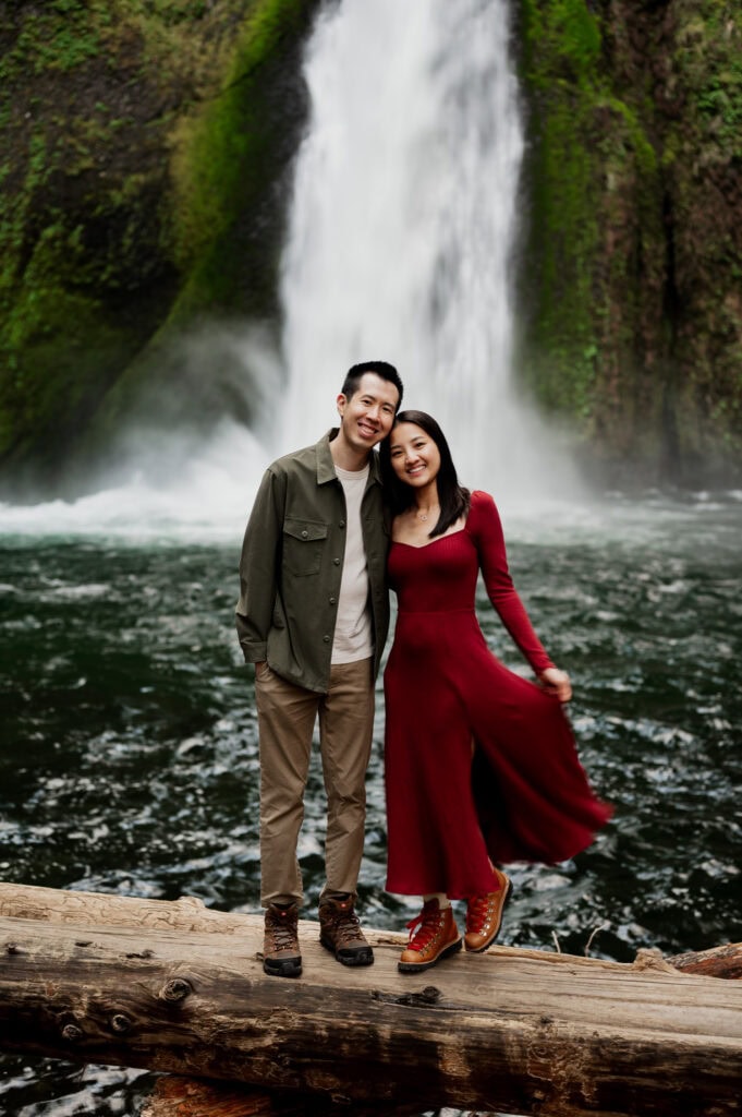 A woman in a red dress and hiking boots stands smiling on a weathered log with her fiancé in a green dress shirt and tan pants in front of a large waterfall in the Columbia River Gorge, photographed by Portland, Oregon wedding and elopement photographer Kyle Carnes.