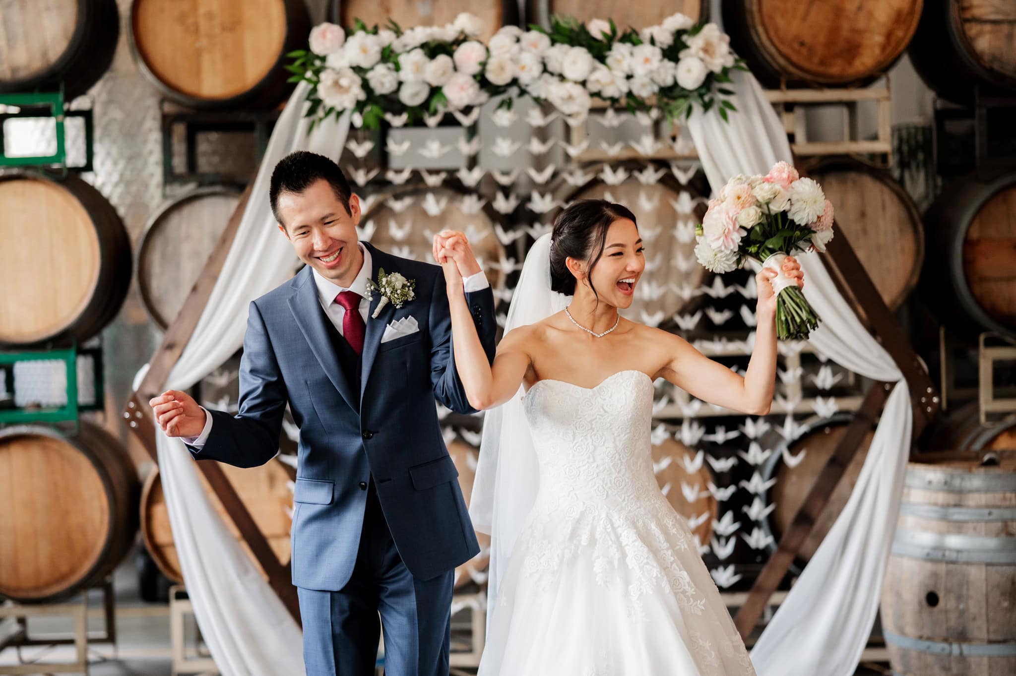 A joyful bride and groom celebrate in a winery setting, surrounded by barrels and floral decorations at Coopers Hall, captured by photographer Kyle Carnes.