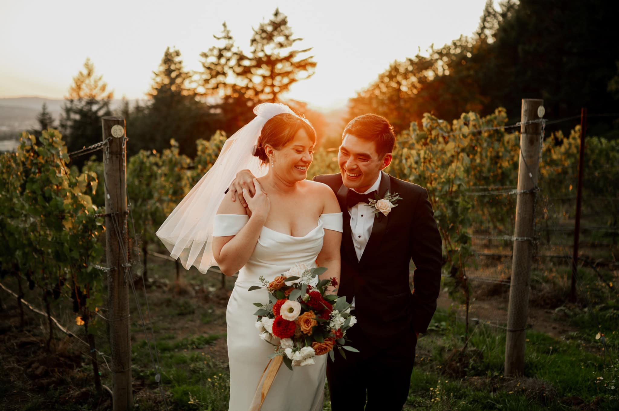 In the warm light of a Portland sunset, the bride and groom smile at each other while standing between lush vineyard rows at Amaterra Winery. The bride holds a bouquet that matches their formal attire.