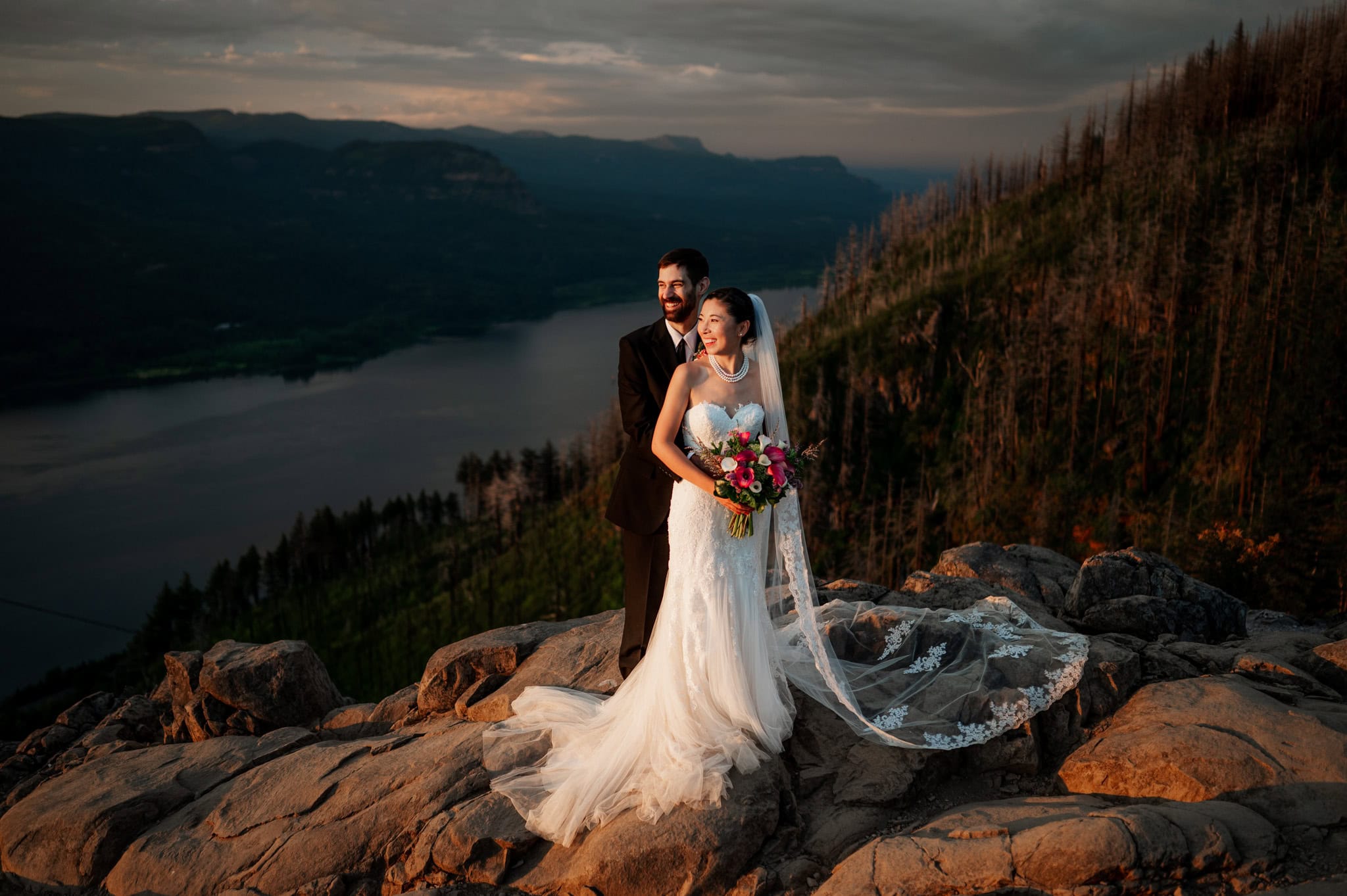 Bride and groom stand on a rocky cliff at Angel's Rest in Oregon's Columbia River Gorge, overlooking a the Columbia River, mountains, and trees. Photographer Kyle Carnes captures the moment.