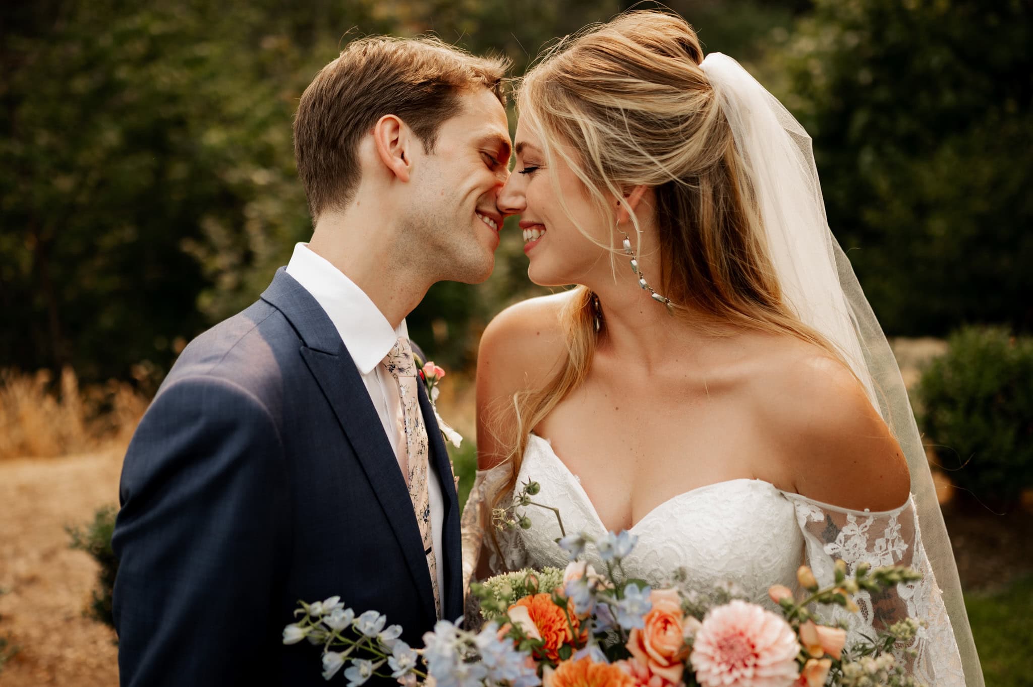 A blonde bride with veil holding a lush bouquet and groom with blue suit and floral tie gently touch noses and smile during their wedding portraits at Cape Horn Estate.