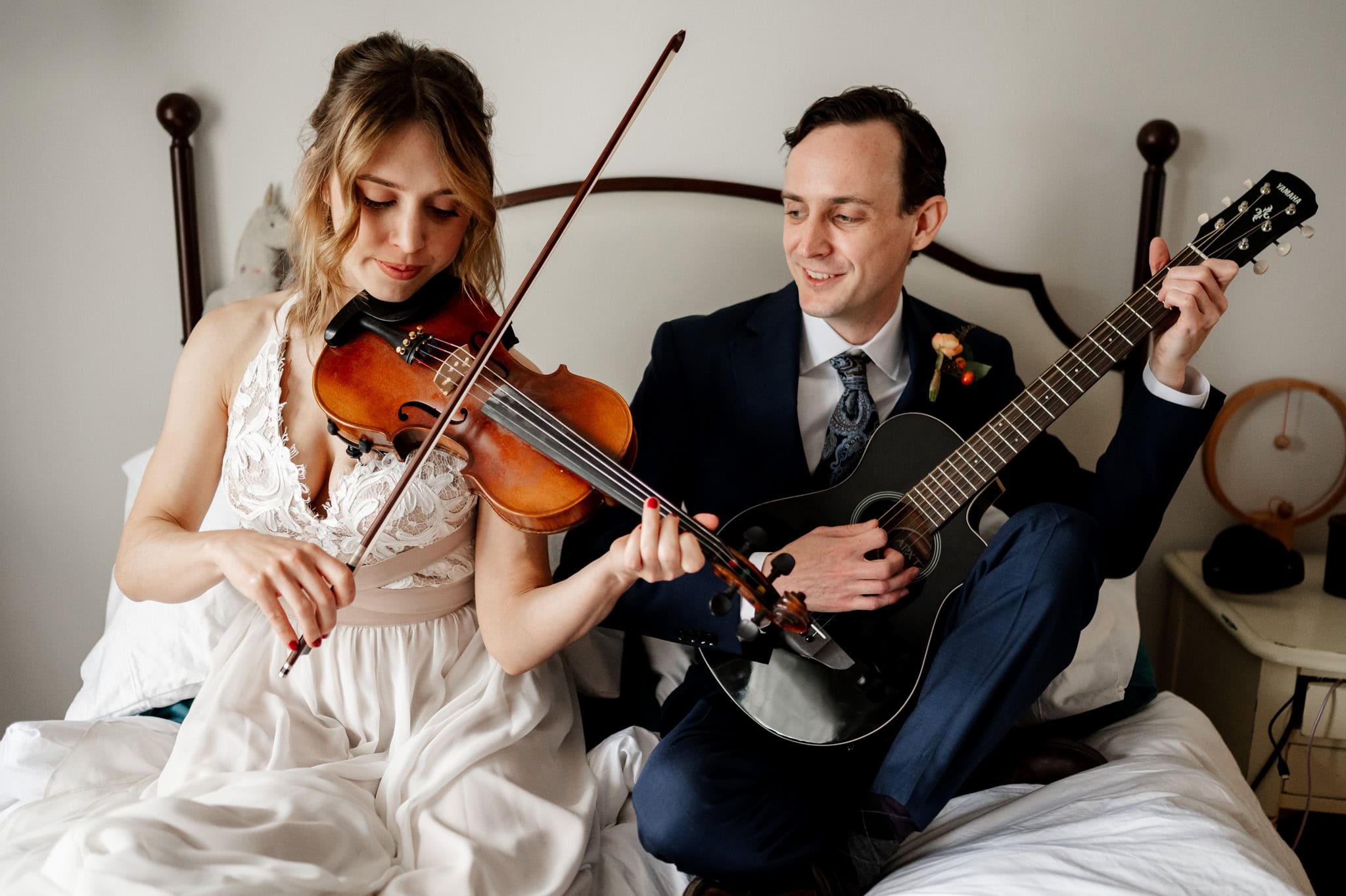 A bride wearing a formal dress plays the violin while seated on a bed next to a groom in formal wear playing the guitar. The setting is intimate, and both are engaged in their music performance.