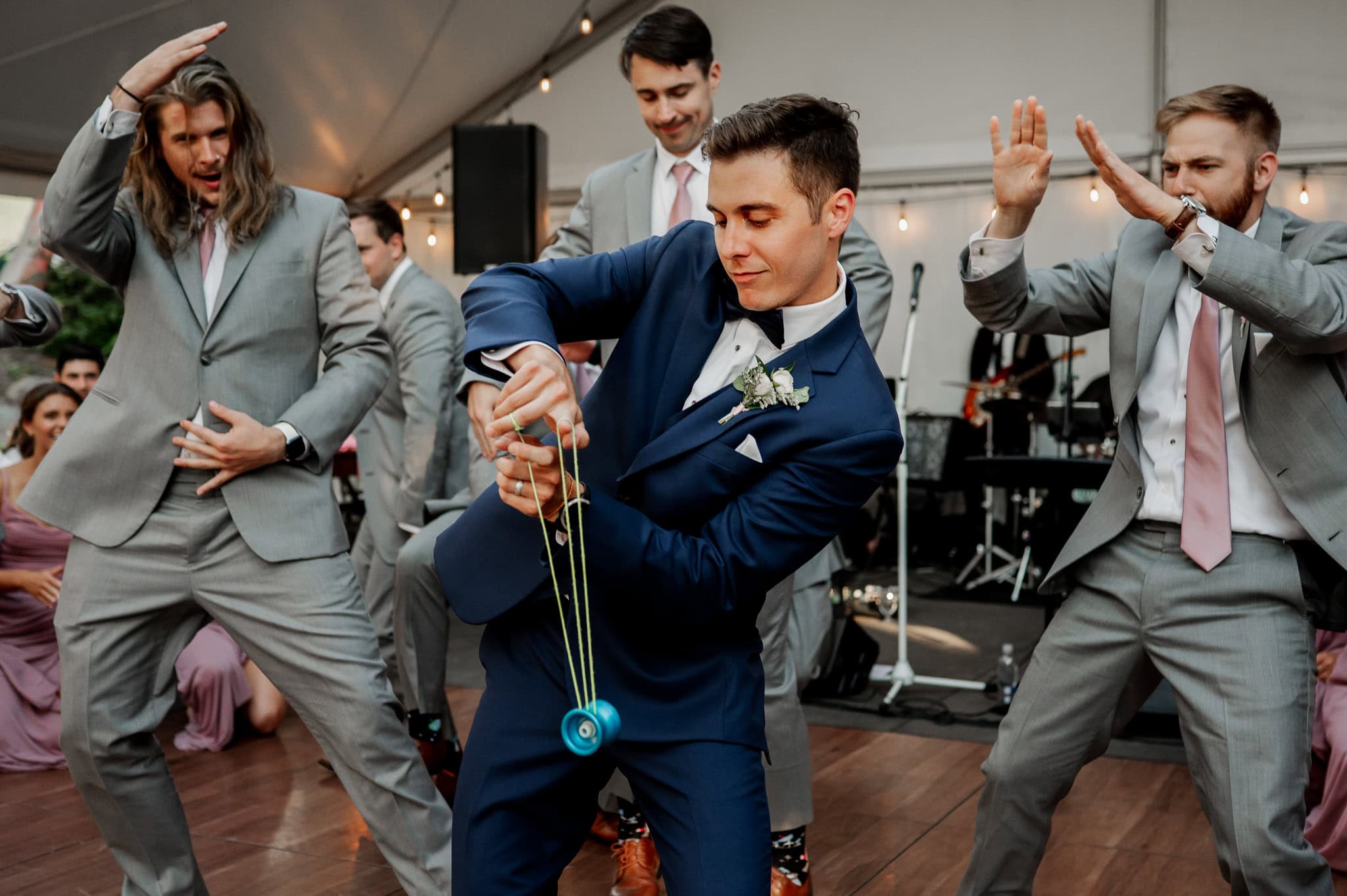 In the middle of a dance floor, a groom wearing a blue suit performs yo-yo tricks as groomsmen in gray suits dance around him at a wedding reception. The scene is captured by wedding and elopement photographer Kyle Carnes in Portland, Oregon.