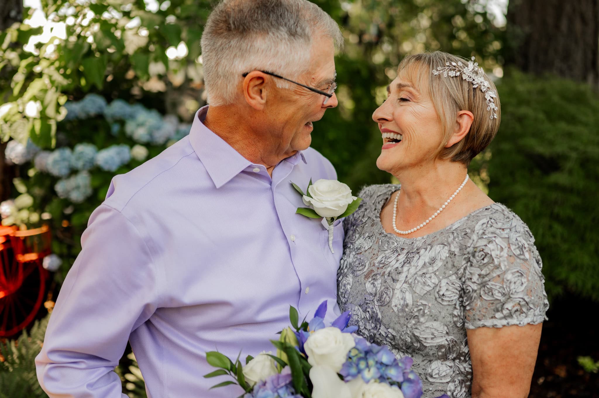 An elderly couple smiles at each other while holding a bouquet in their lush garden. They are captured in an intimate moment near Portland by Oregon wedding and elopement photographer Kyle Carnes.