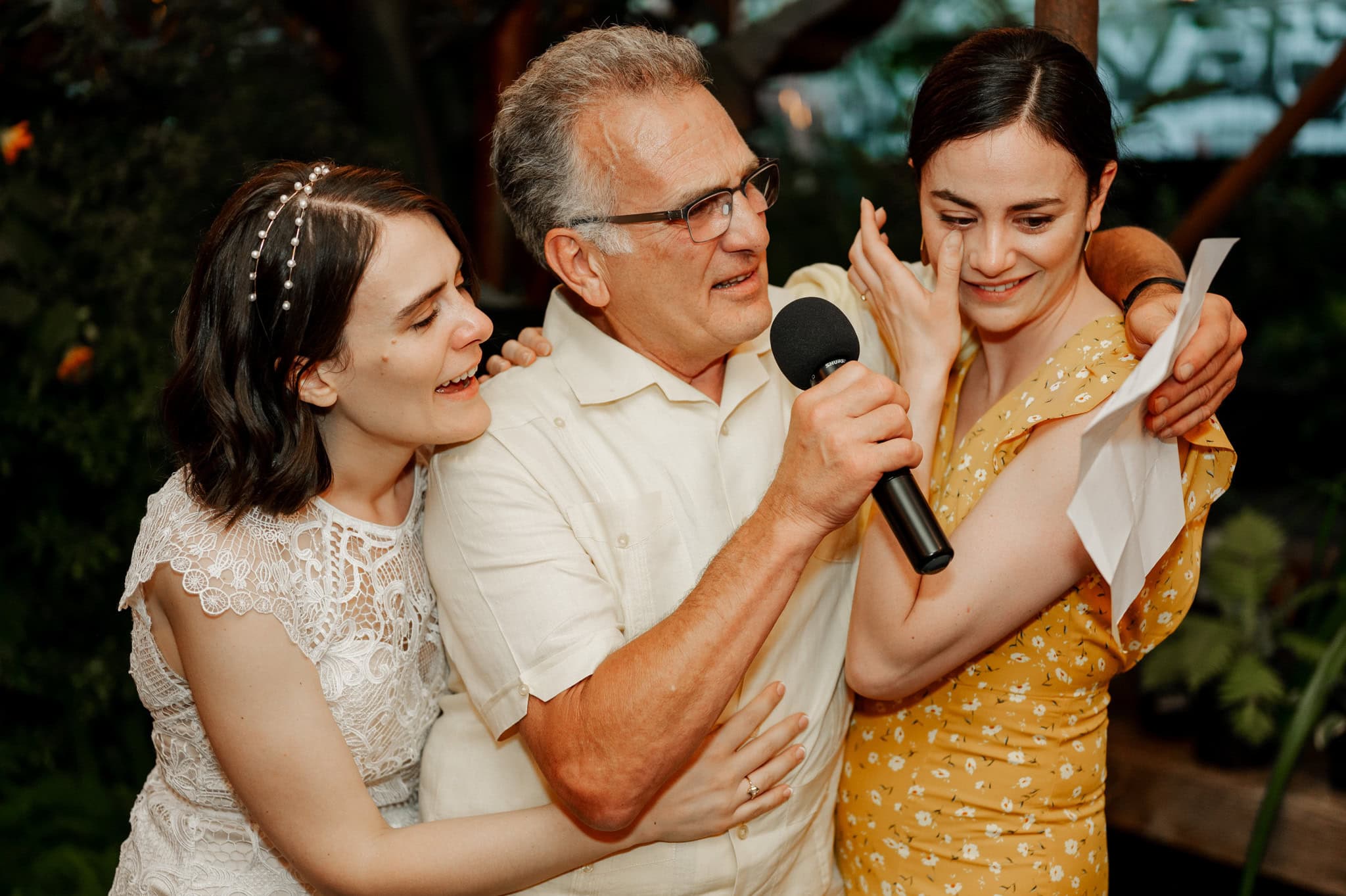 The father of the bride gives a speech, holding a microphone and reading from a piece of paper, the bride in a white dress and her sister in a yellow floral dress embrace him warmly. This moment was captured at a wedding by Oregon wedding and elopement photographer, Kyle Carnes, at Blockhouse in Portland.