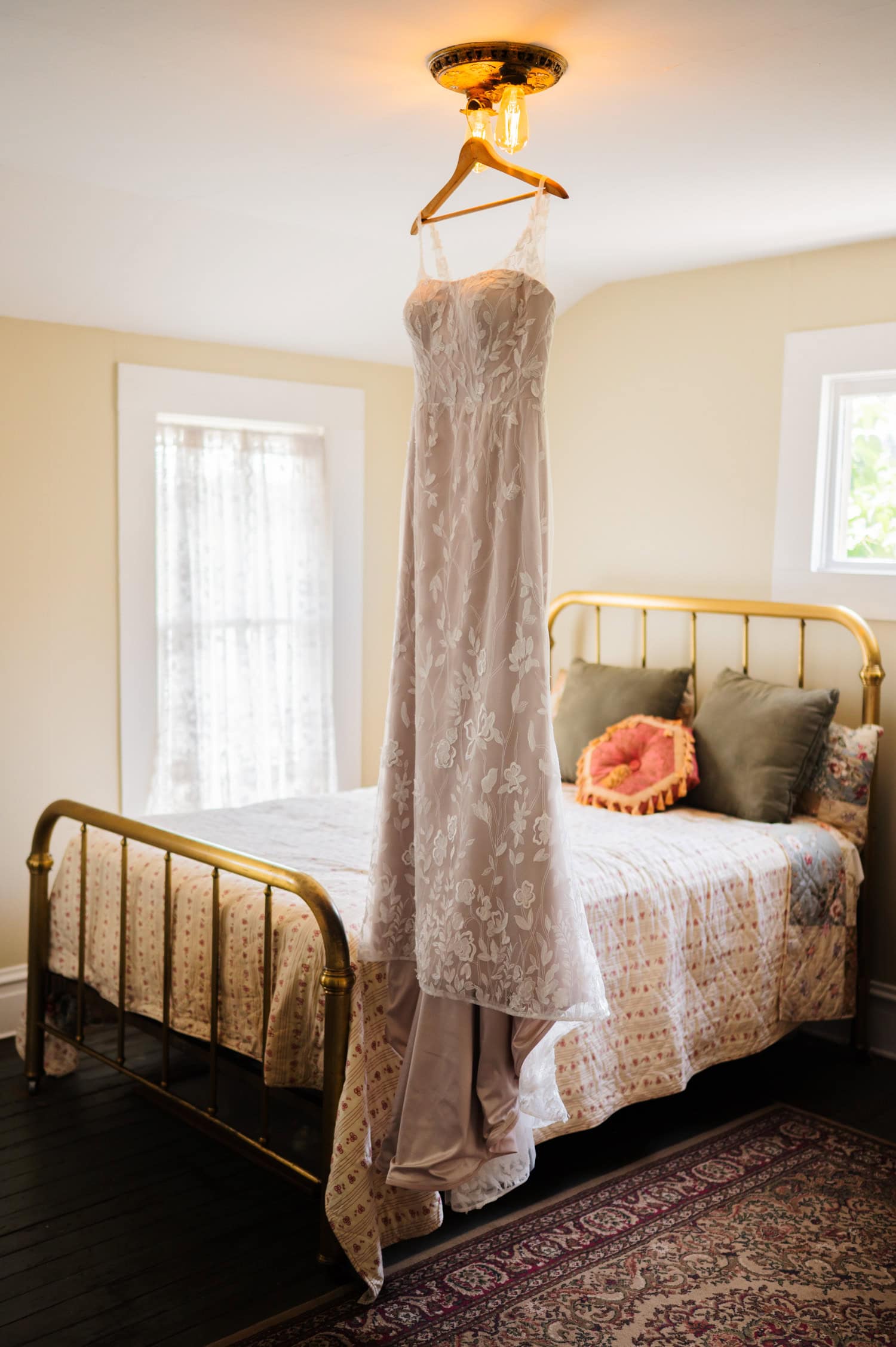 A strapless white wedding dress hangs elegantly on a light fixture above a bed in a cozy room, awaiting the Rossi Farms wedding photos.