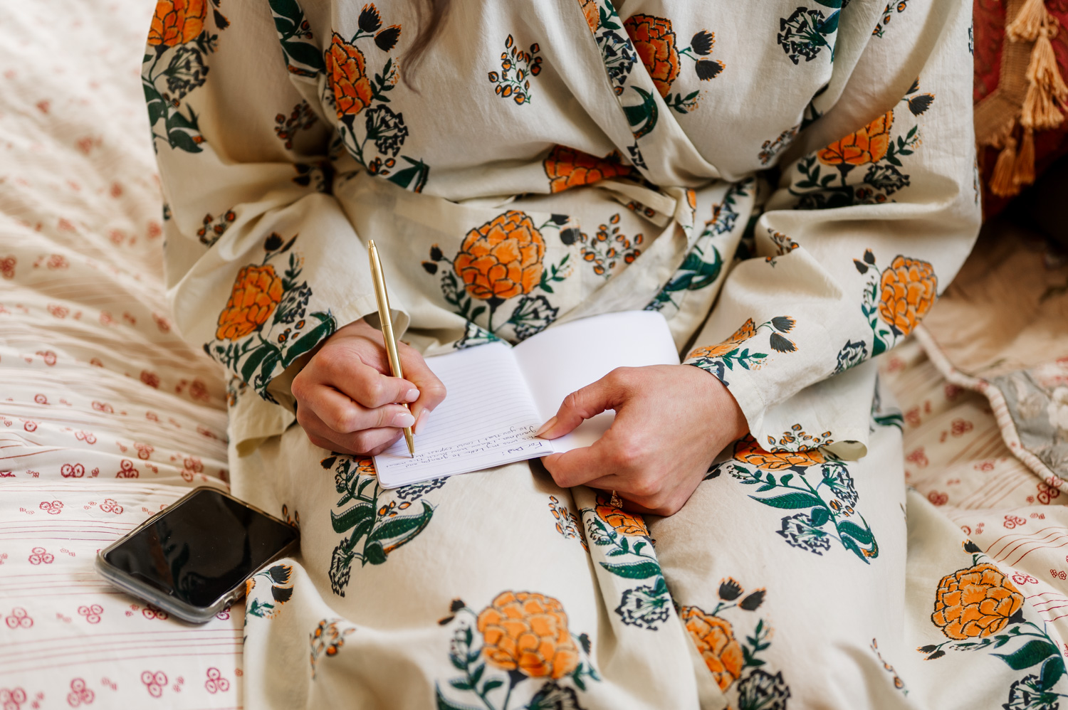 A person wearing a floral robe sits on the bed writing in a notebook with a pen. A phone is placed nearby, and the scene conveys a peaceful moment before sharing wedding photos from Rossi Farms.