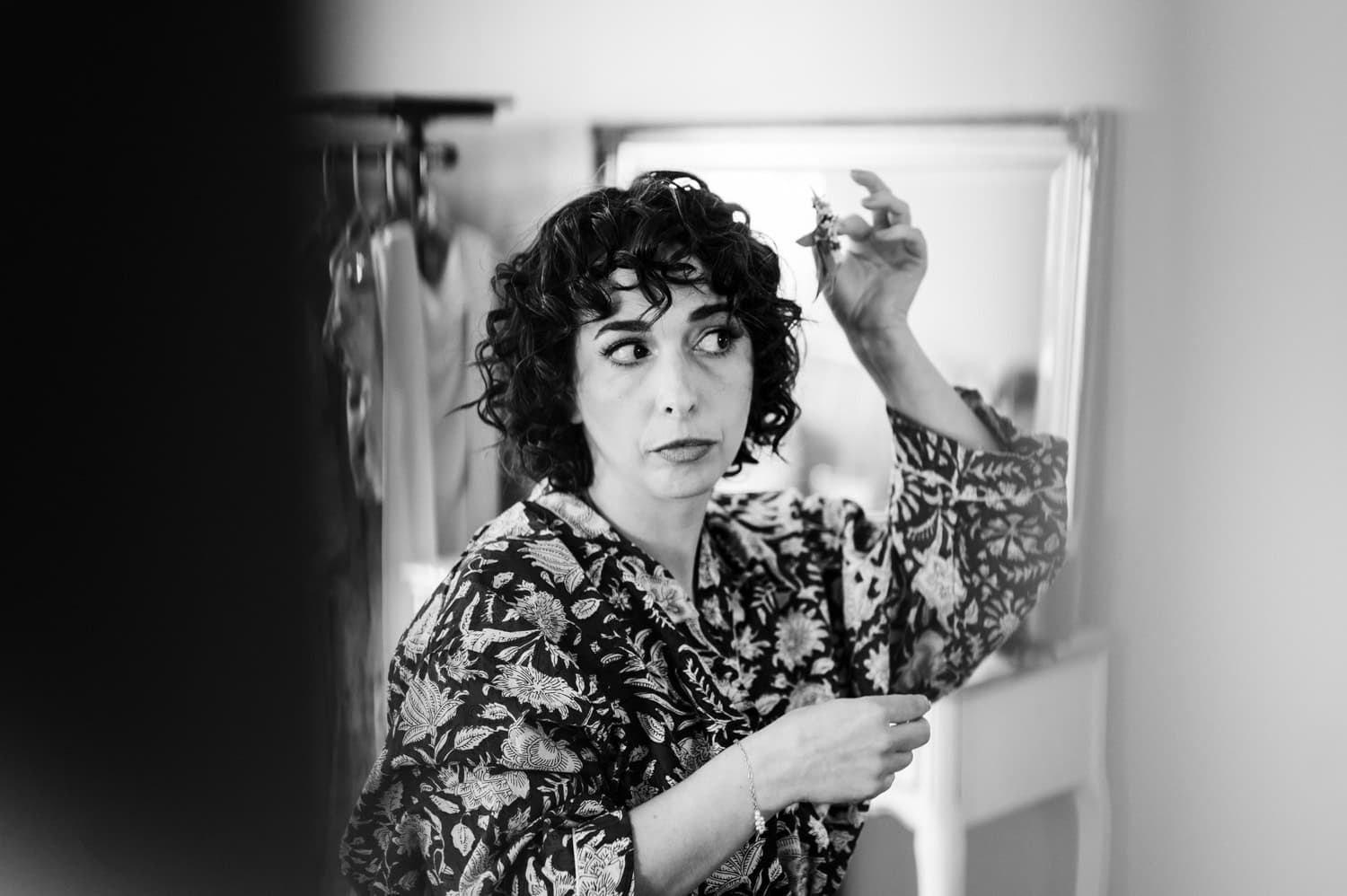 A woman with curly hair looks into a mirror while adjusting her curls, wearing a patterned top. The black and white photo captures her timeless beauty and style.