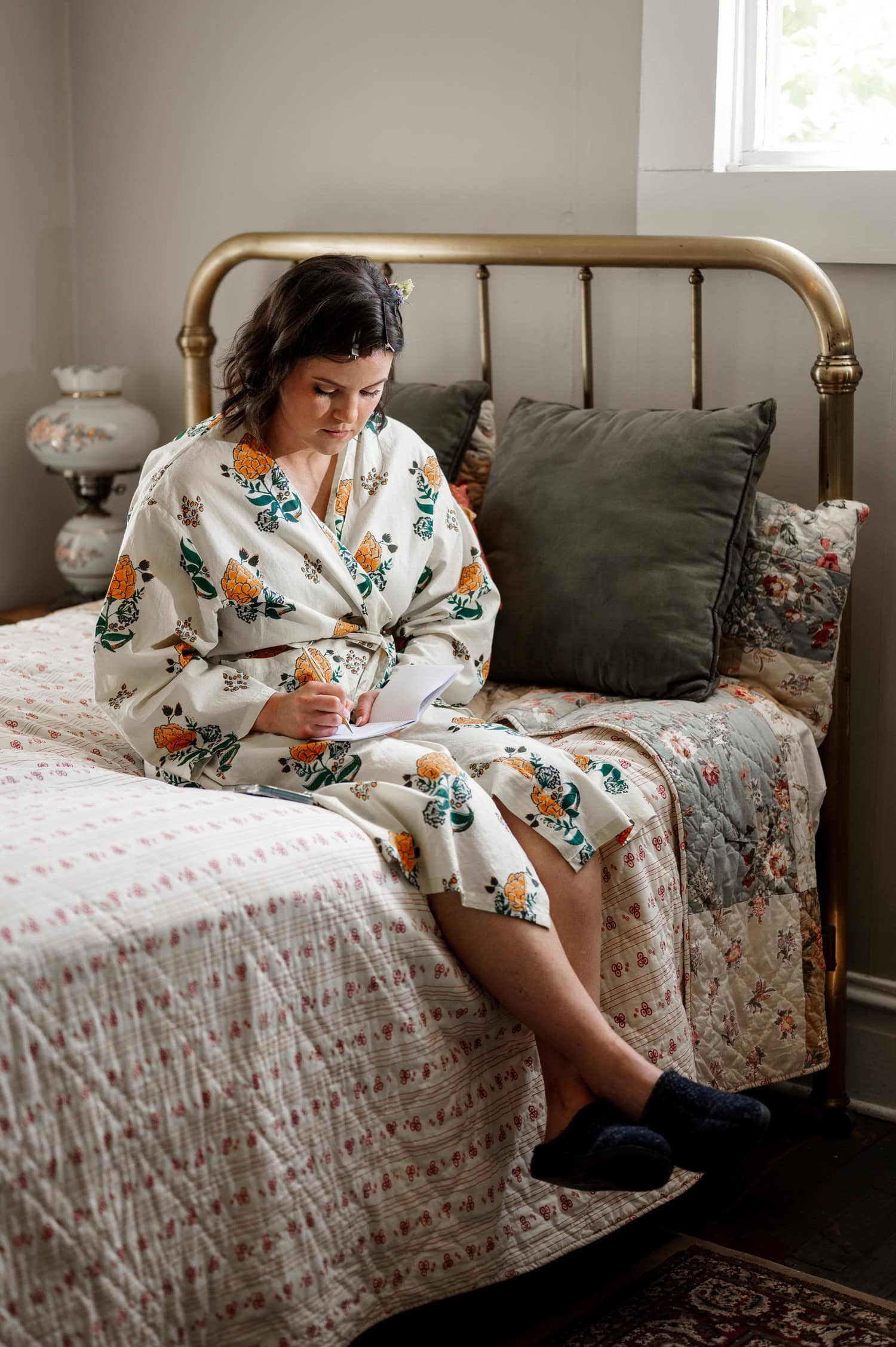 Person in a floral robe sits on the bed by the window, writing in a notebook. Wedding photos from Rossi Farms are hung on the wall, reflecting softly.