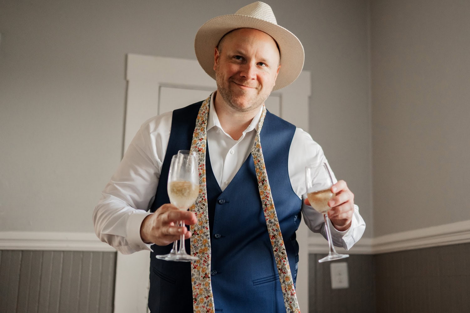 A smiling man in a blue vest and hat holds two glasses of champagne, conveying a joyful ambiance associated with Rossi Farms wedding celebrations.