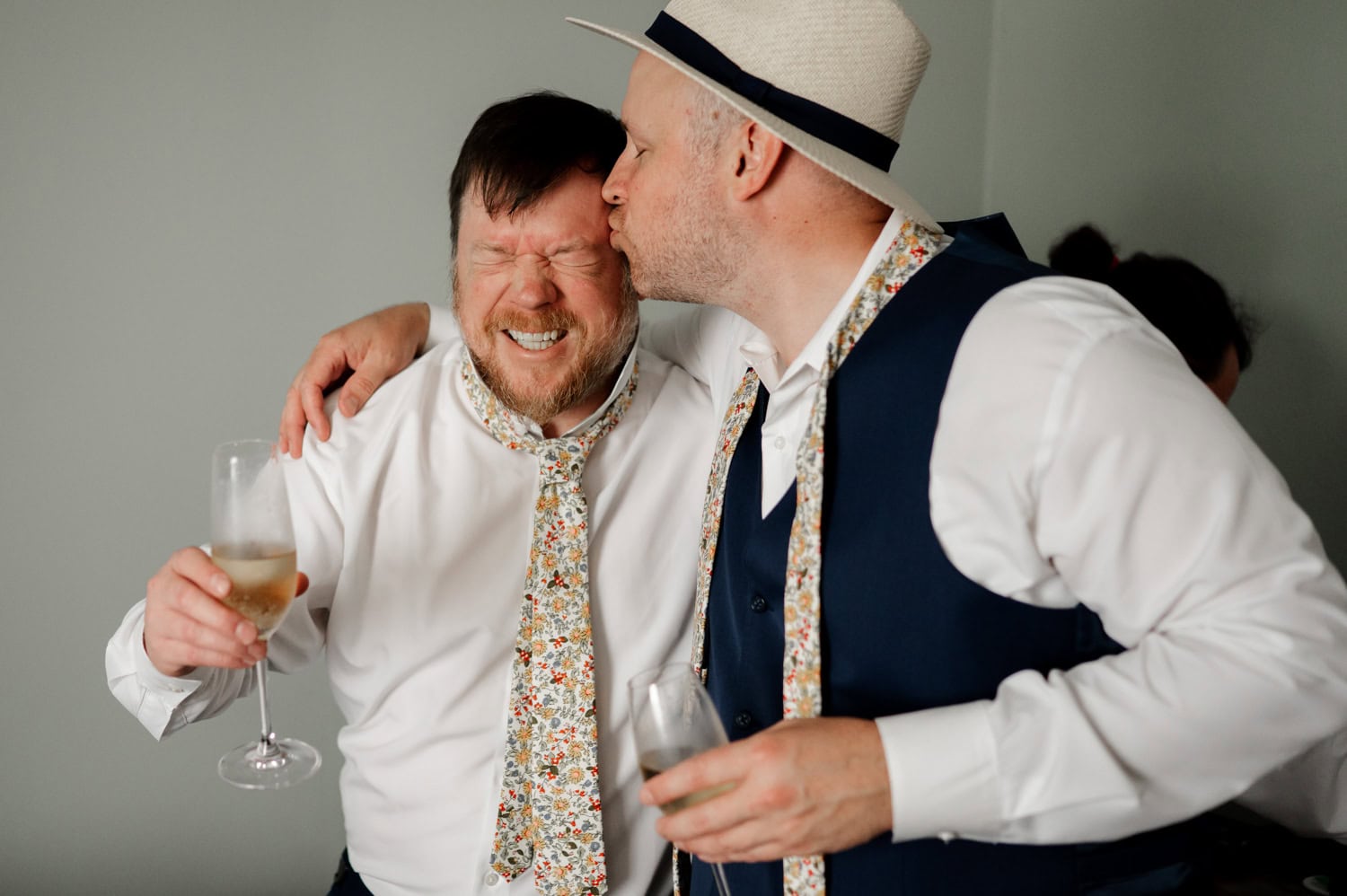 Two men with matching floral ties share a joyful moment at Rossi Farms wedding. One kisses the others cheek while both smile and hold drinks, embodying love and celebration.