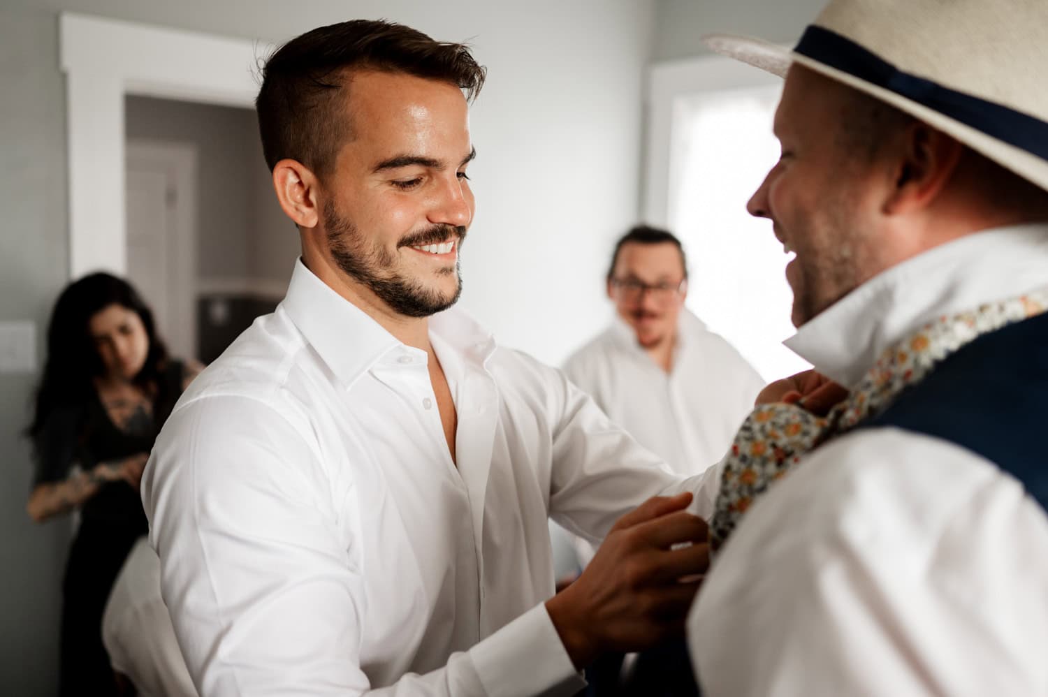 A man helps another with his tie while two others stand in the background, all smiling in a cheerful indoor setting. The group is dressed casually and appears to be enjoying the moment together.