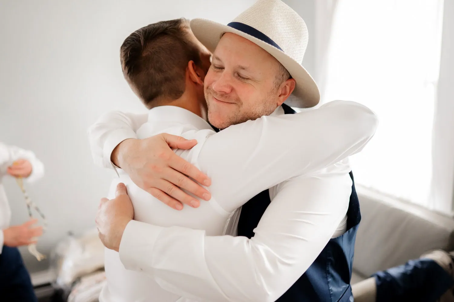 Two men in formal attire warmly embrace in a well-lit room at Rossi Farms. One wears a hat and both display happy expressions, capturing the joy of the moment.