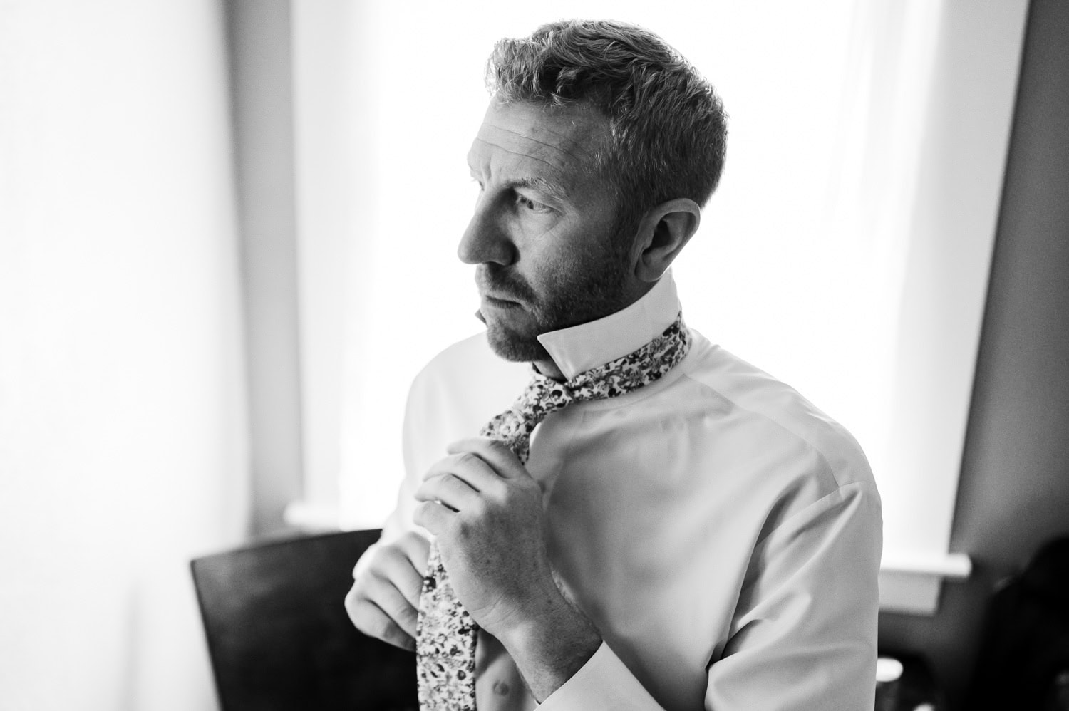 Man in a white shirt adjusting a floral tie, looking thoughtful at the Rossi Farms wedding. Black and white image with soft natural light illuminating his face.