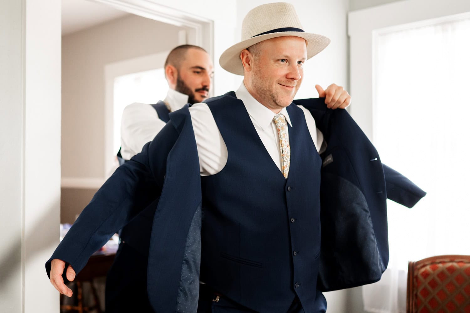 A man in a hat gets help putting on his suit jacket in a well-lit room, getting ready for wedding photos at Rossi Farms.