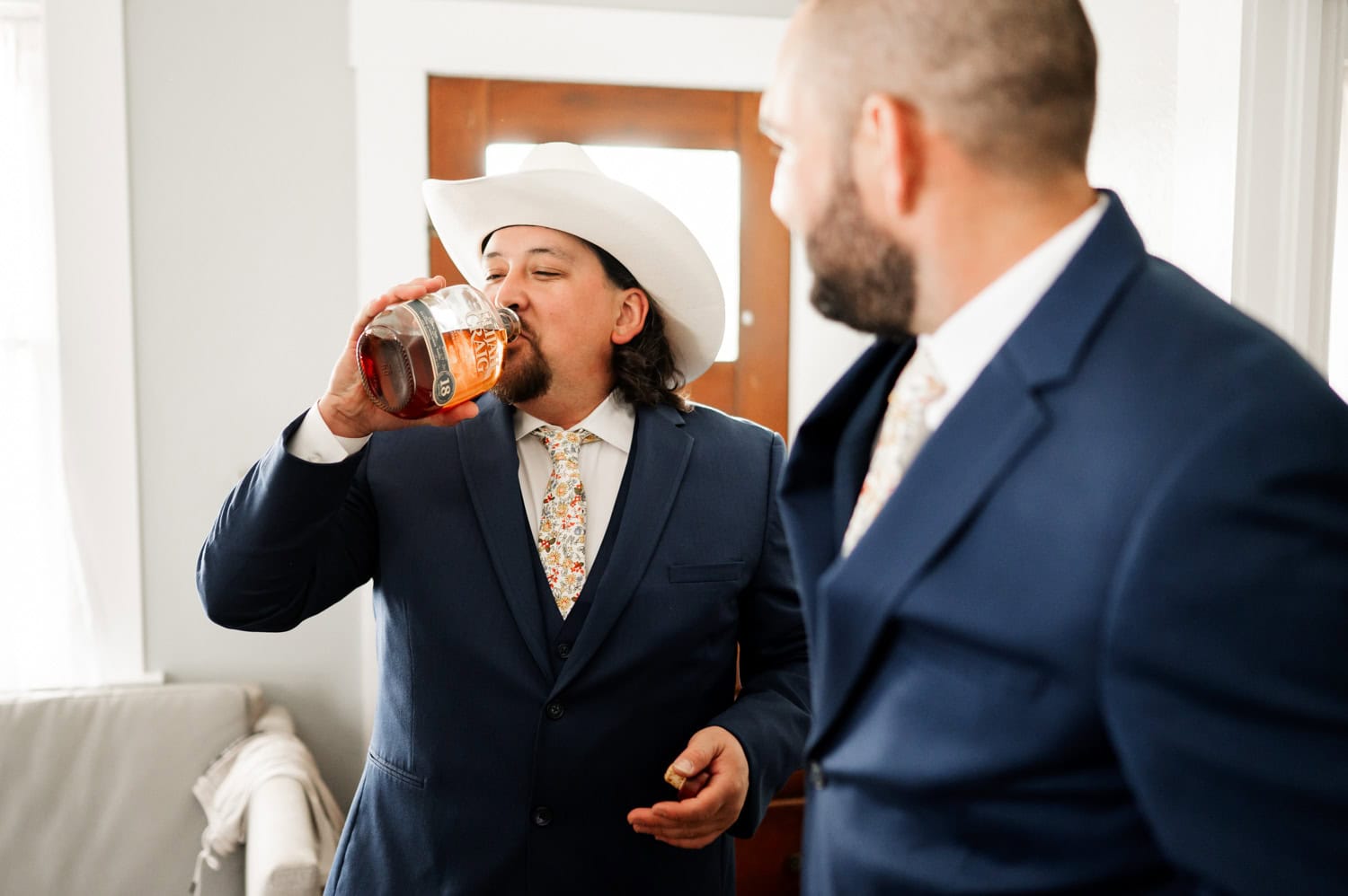 A man in a suit and cowboy hat takes a drink from his bottle as another man in a suit watches with an amused expression at a Rossi Farms wedding.