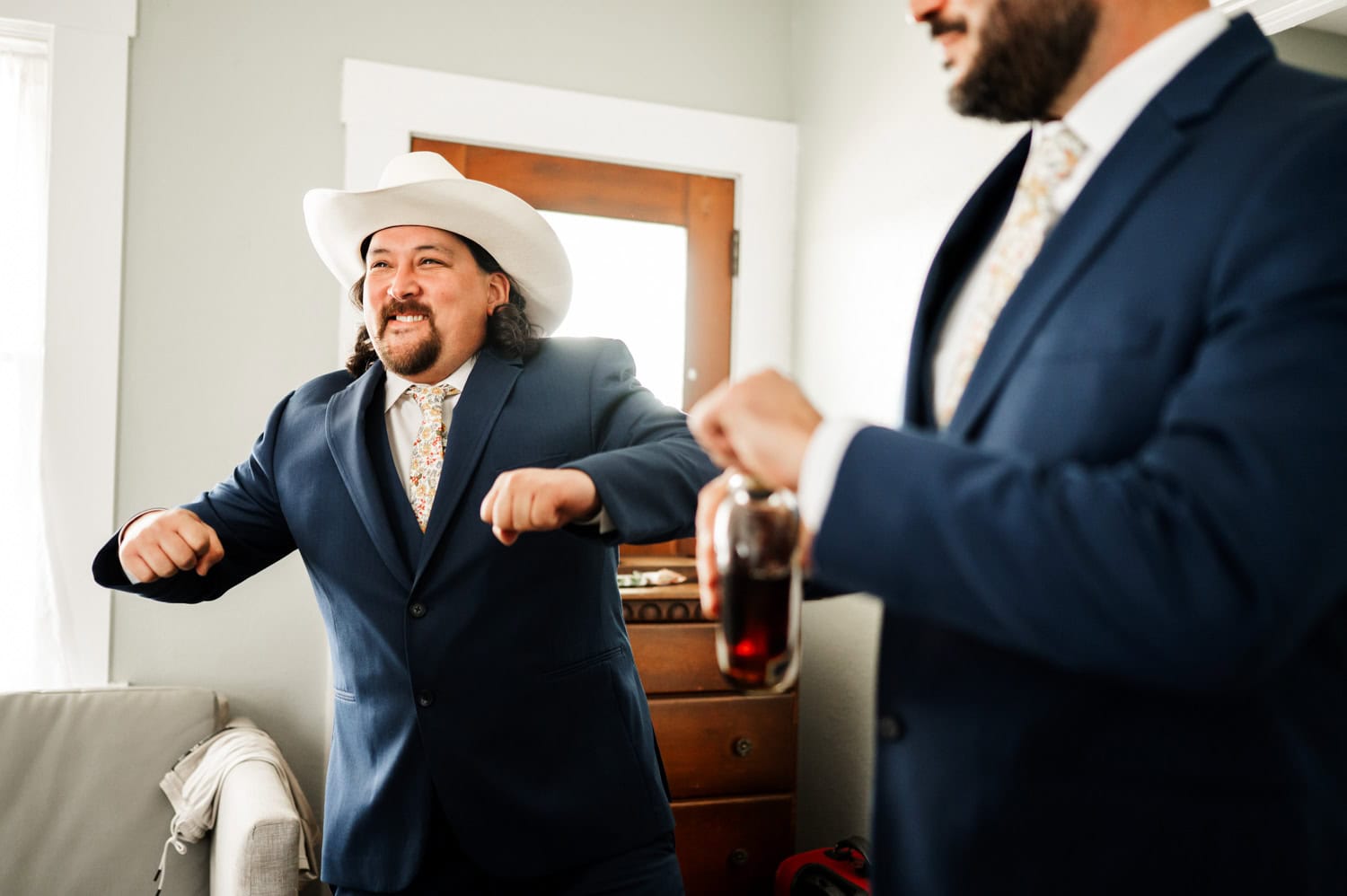 Man in cowboy hat and suit smiling enthusiastically, standing beside another man in a suit who is holding a drink at Rossi Farms. Both have lively expressions, conveying joy and camaraderie at a wedding event.
