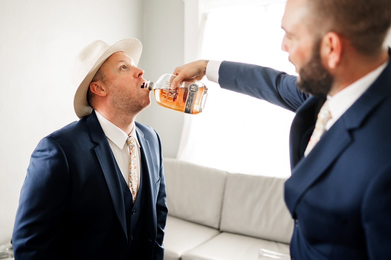 In a bright room, a man in a suit pours whiskey into another mans mouth, capturing a moment of celebration and camaraderie.
