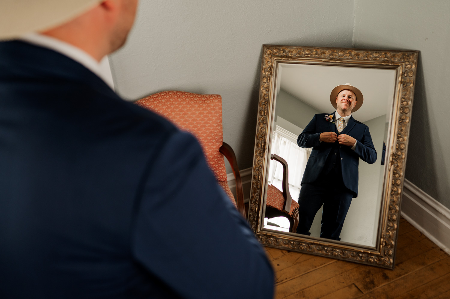 Man in suit and hat adjusting clothes, smiling at himself in a standing mirror, preparing for wedding photos at Rossi Farms.