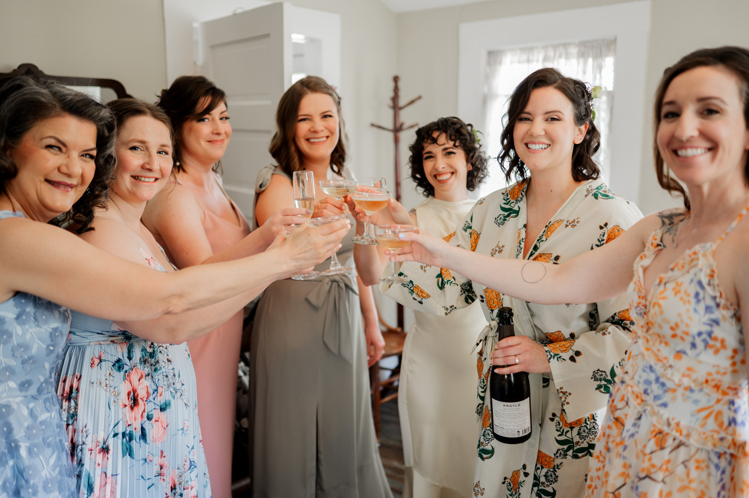 Seven women in dresses raise their champagne glasses in a toast, smiling and celebrating indoors.