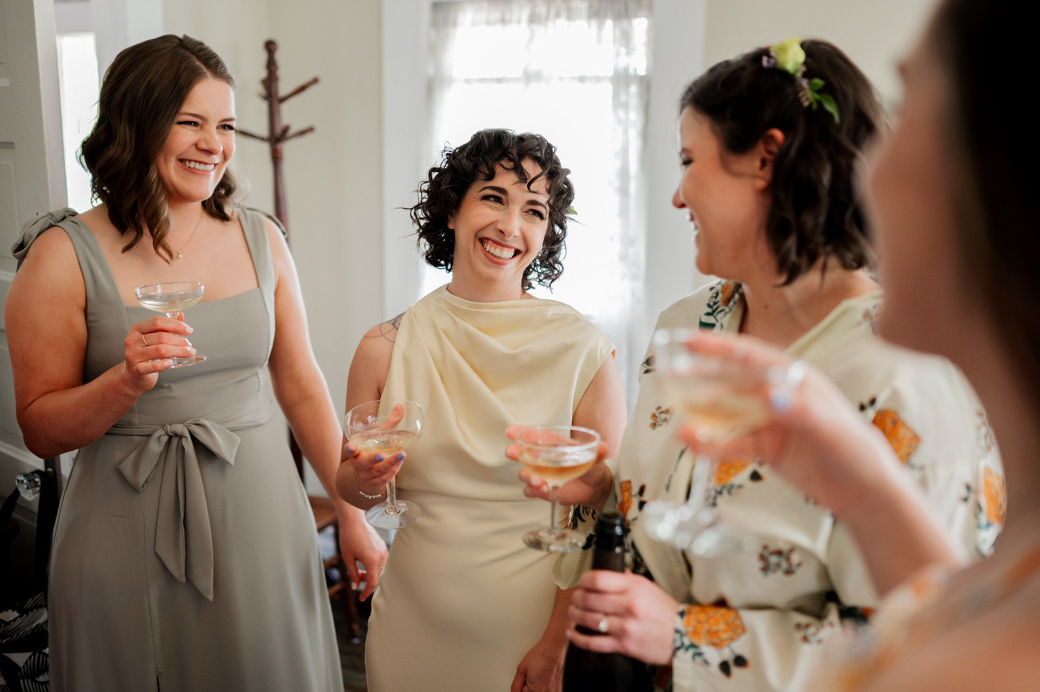 Four women elegantly dressed, smiling with drinks in hand, gather in a warmly lit room.