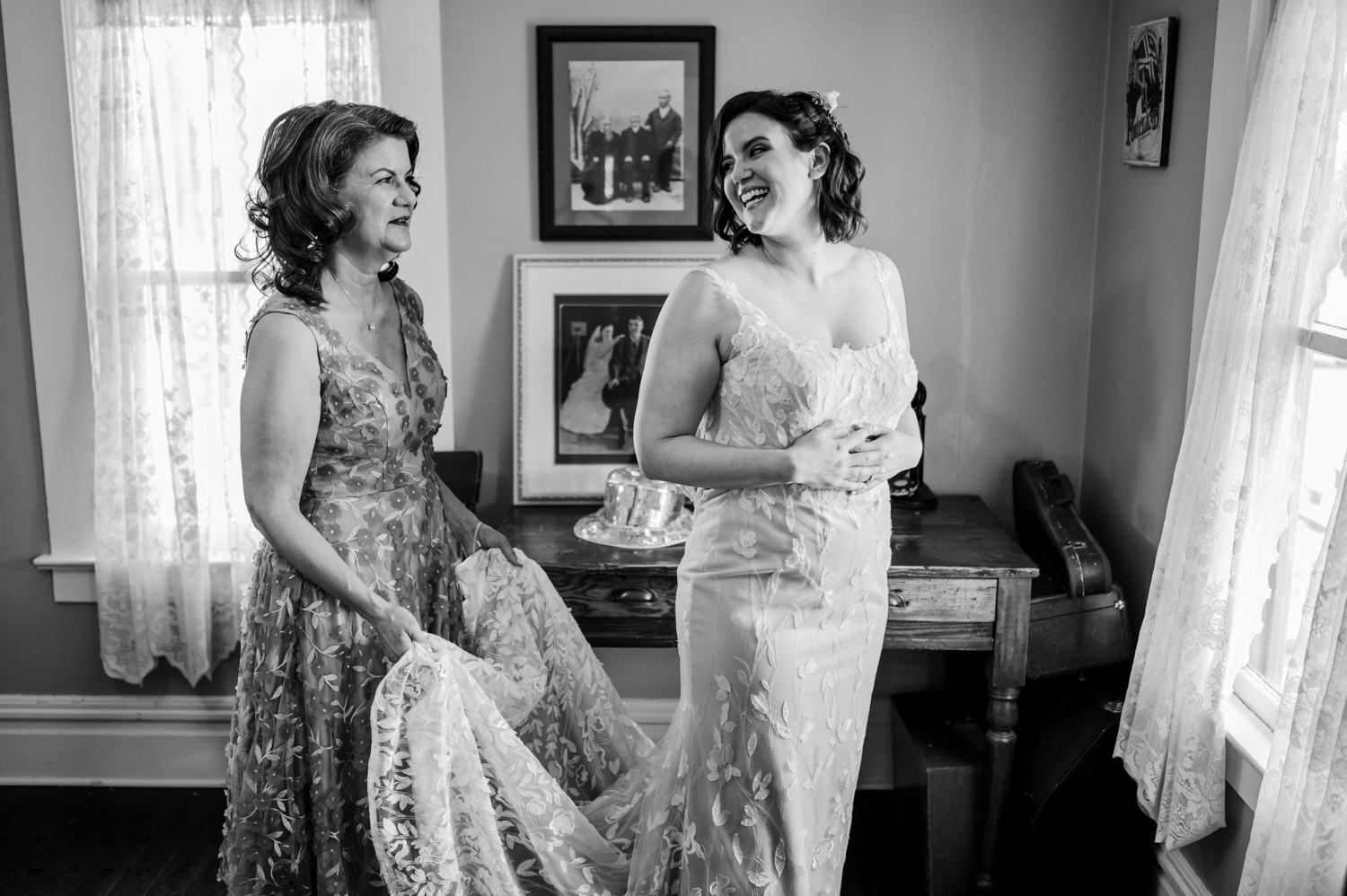 Bride and another woman laughing together in a room with lace curtains and vintage photos, capturing a joyful moment at Rossi Farms.