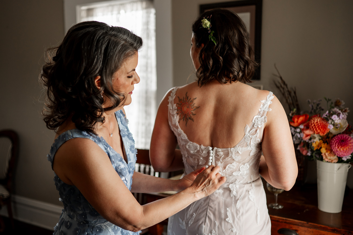 A woman in a blue dress helps another woman with the buttons on her wedding gown, revealing a sunflower tattoo on her back.