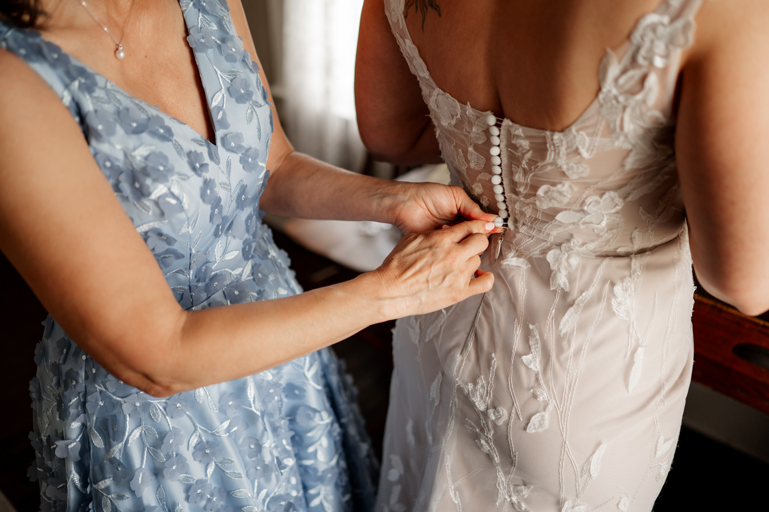 At a Rossi Farms wedding, a person in a blue floral dress assists another with buttoning their lace wedding gown.