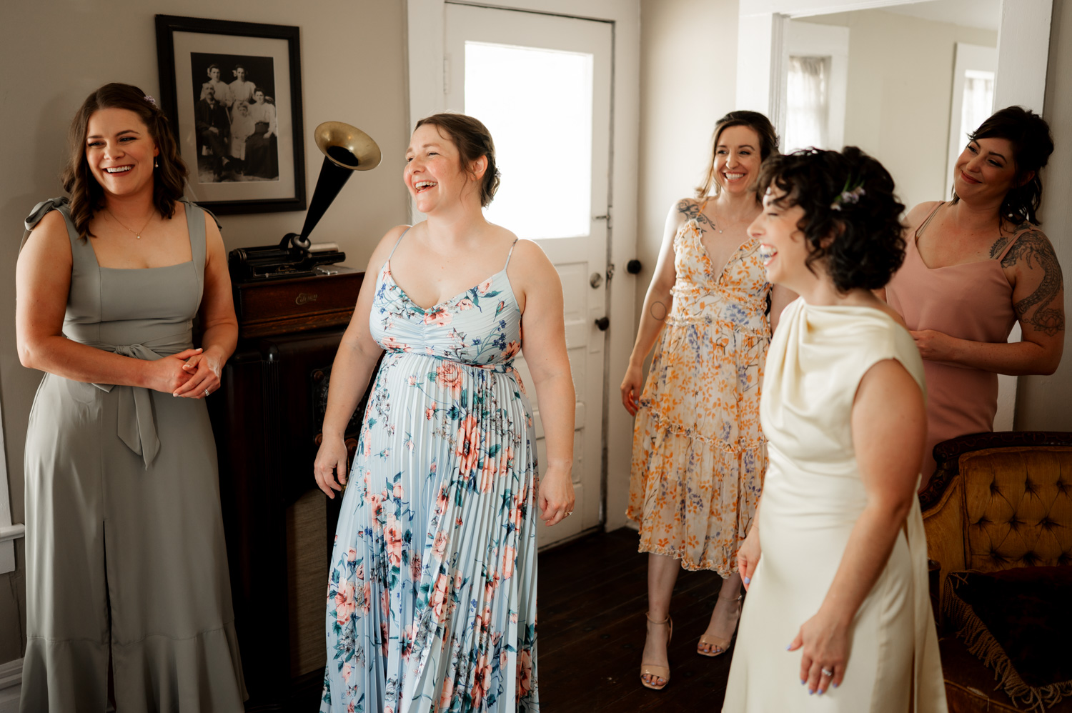 Five women in dresses laugh and smile while standing together, surrounded by vintage decor at a Rossi Farms wedding.