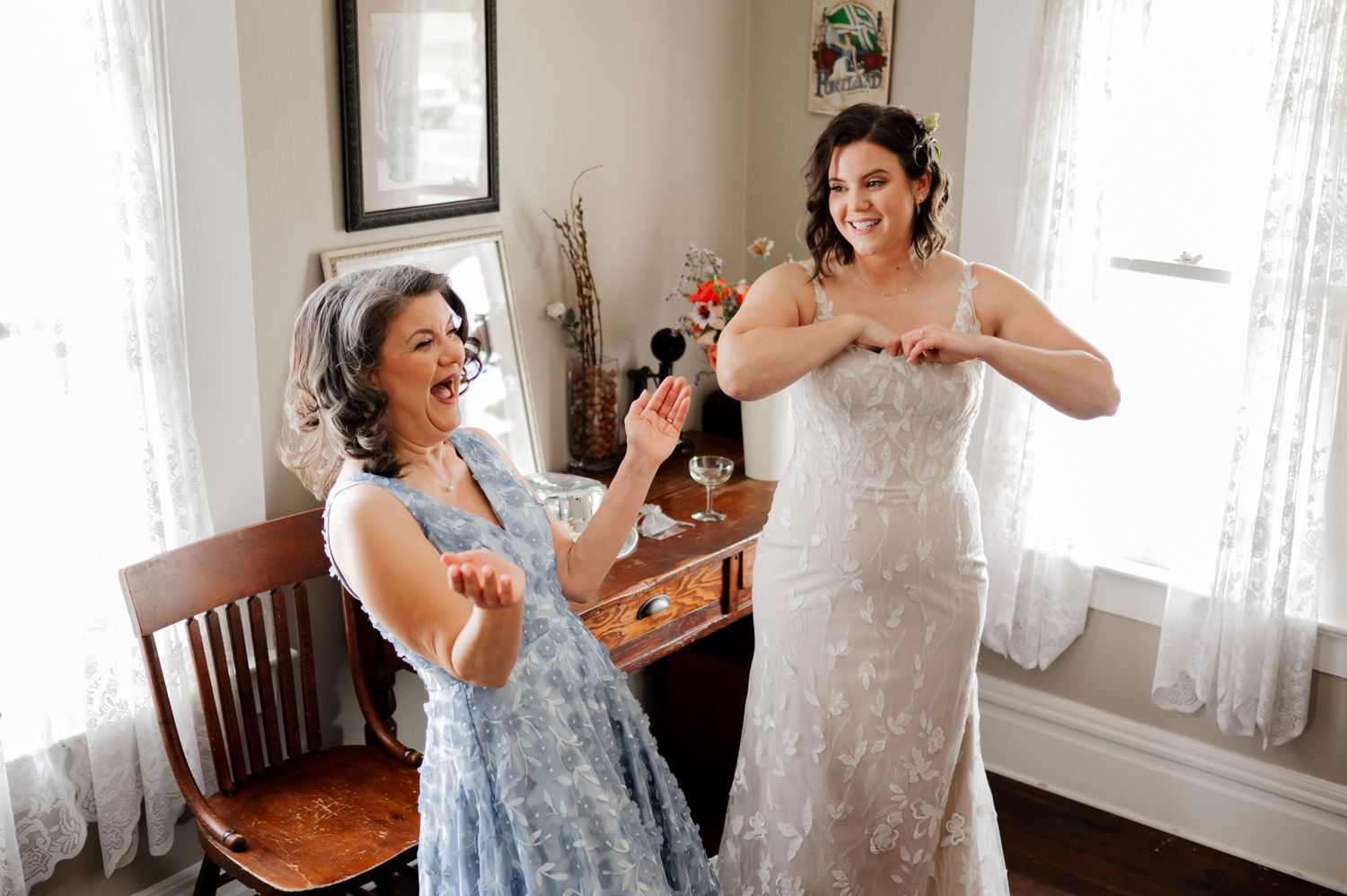 In a sunlit room with vintage decor, a bride in a white dress and a woman in blue dresses enjoy a joyful moment together, captured in wedding photos at Rossi Farms.