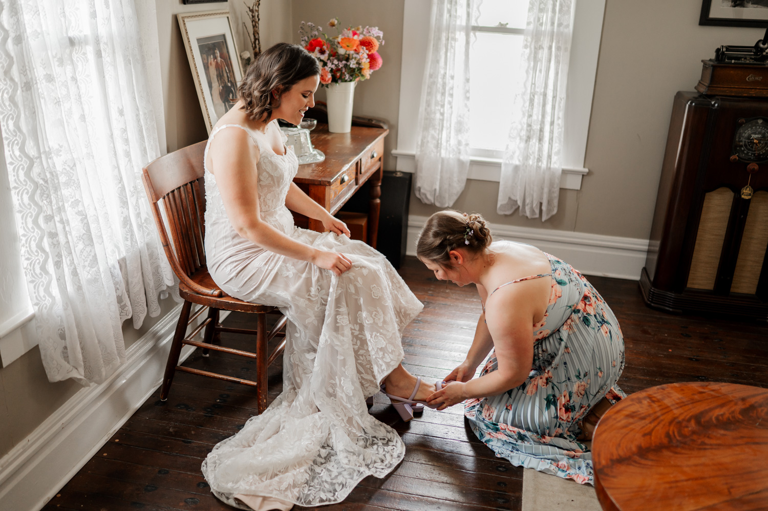 At a sunlit Rossi Farms wedding, the bride in an elegant white dress sits on a chair as her friend kneels next to her, adjusting the brides shoes.
